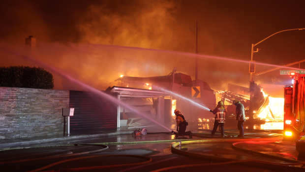 A nighttime image of CAL FIRE crews battling a raging fire in Pacific Palisades. Firefighters spray water from multiple hoses at a burning structure, with flames and thick smoke consuming the background. The intense orange glow of the fire lights up the scene, while firetrucks with flashing emergency lights stand nearby. A stone wall and a partially burned structure are visible in the foreground.