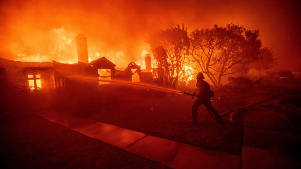 A firefighter sprays water on a burning house engulfed in flames during a wildfire, with intense orange and red hues lighting up the night sky. Nearby trees and structures are silhouetted against the blaze, capturing the urgency and devastation of the scene.