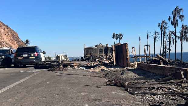 A photo of the aftermath of the Palisades Fire along the Pacific Coast Highway, showing two Los Angeles County Sheriff’s vehicles parked near the burned remains of buildings and palm trees. A clear blue sky and ocean are visible in the background, contrasting with the destruction.