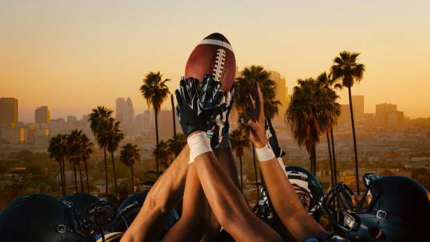 A group of football players wearing helmets and gloves raise a football together against a backdrop of palm trees and the Los Angeles skyline at sunset. The image captures a celebratory and competitive spirit, symbolizing the excitement of Super Bowl 2025 in L.A.
