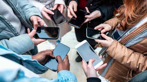 A group of students standing in a circle, each holding a smartphone with blank screens. The image highlights the prevalence of mobile device usage among young people, a key issue behind the LAUSD’s recent decision to ban cellphones in classrooms.