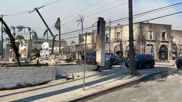 A scene of devastation from recent wildfires shows burned-out buildings, charred palm trees, and downed power lines. Vehicles and emergency response teams are present amid the wreckage, highlighting the aftermath of the disaster.