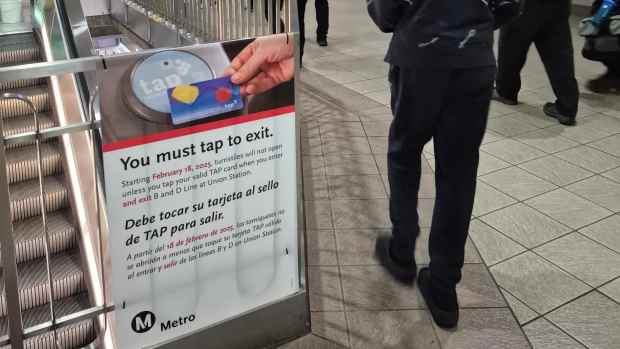 A sign at an LA Metro station informing passengers about the “Tap to Exit” program, requiring TAP card validation to exit starting February 18, 2025. In the background, commuters walk through the station, some carrying backpacks, while Metro security personnel monitor the area. Overhead lights illuminate the modern, tiled interior.