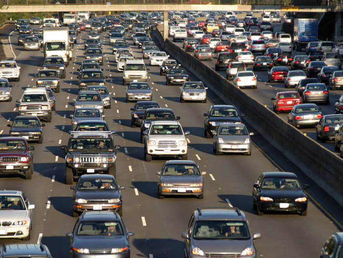 A photo of a multi-lane highway filled with dense traffic in both directions during daylight. Vehicles of various types and colors, including sedans, SUVs, and trucks, are tightly packed, highlighting the intensity of rush hour. A concrete divider separates the lanes, with an overpass visible in the background.